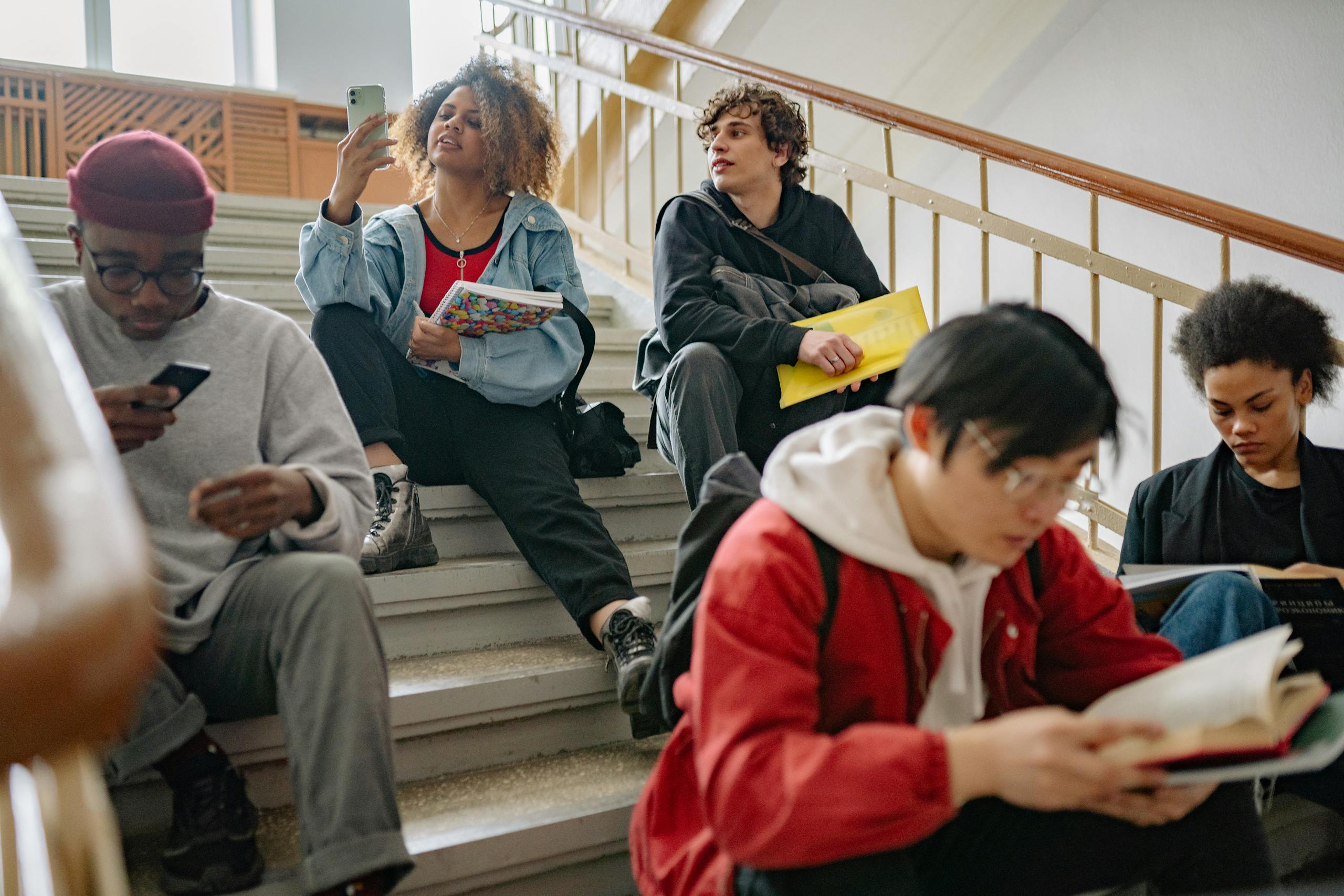 College students sitting on stairs, studying and using phones in a university setting.
